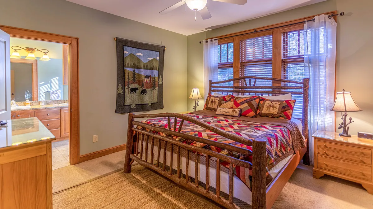 A cozy bedroom with a rustic log bed frame, topped with a colorful quilt. Soft morning light filters through the window, which is accented by a decorative wall hanging and a nightstand with a warm bedside lamp.
