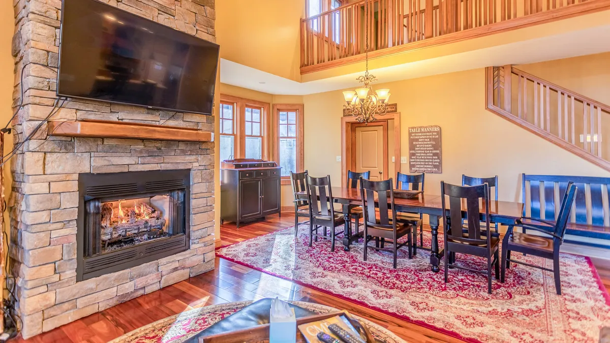 A warm, open dining area features a long wooden table with seating for ten beneath an elegant chandelier. The dining space is seamlessly connected to the great room, with a fireplace visible nearby and large windows letting in an abundance of natural light.