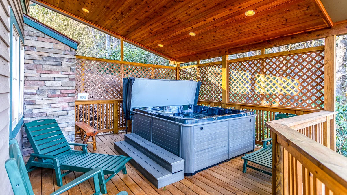 A tranquil exterior deck scene with a private hot tub, green chairs for lounging, and a lattice enclosure offering partial privacy. The setting is surrounded by nature, creating an inviting space for evening soaks or morning coffee.