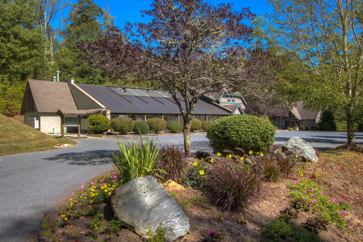 Showcasing a landscaped garden with seasonal flowers, the building's facade, a driveway, and striking rock formations, this image captures the inviting natural beauty of the property and hints at the broader mountain environment beyond.