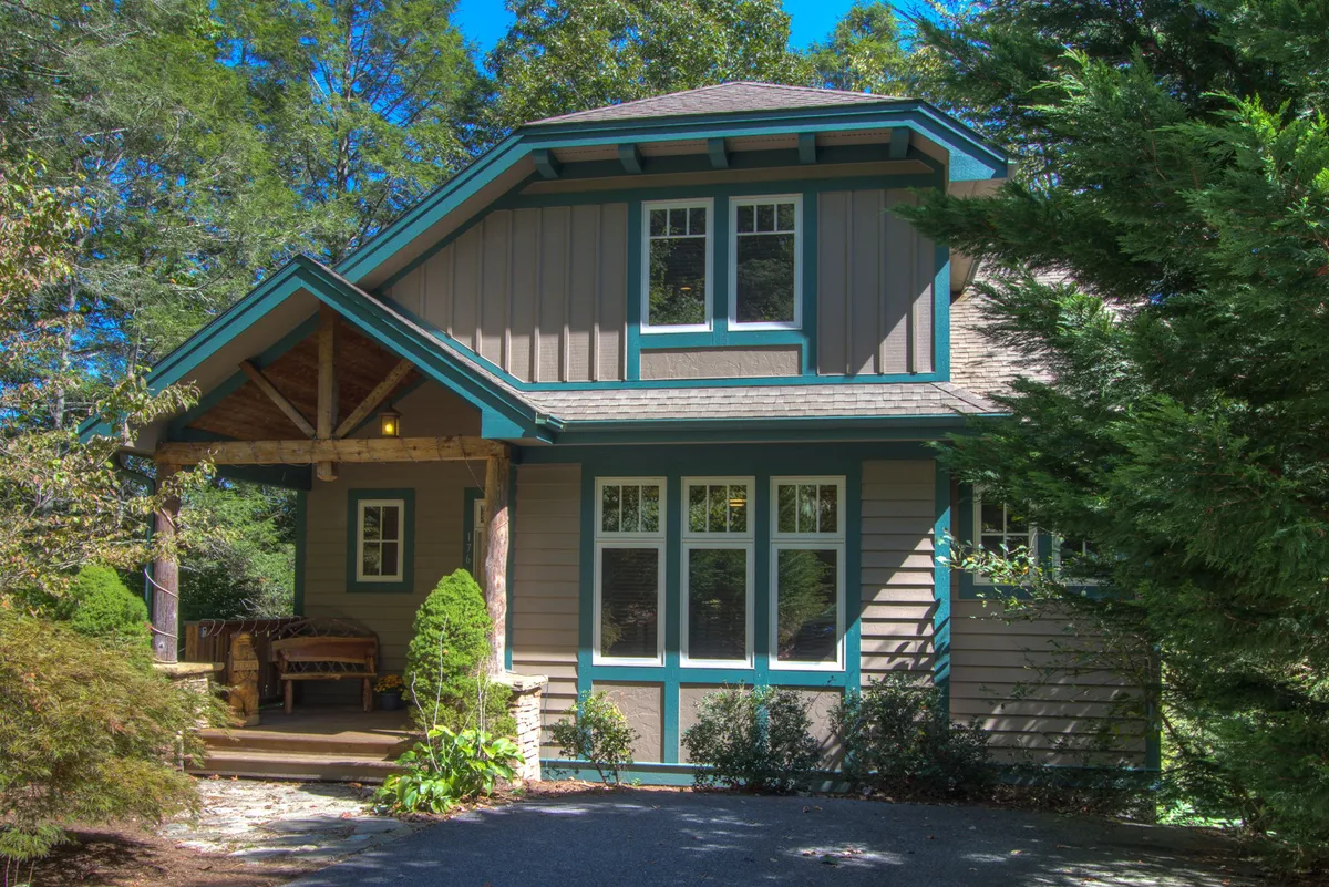 The image showcases the inviting exterior of Bearfoot Lodge, featuring a welcoming front porch with comfortable seating, intricate decorative trim, large windows that promise beautiful natural light, and lush landscaping set against a tranquil mountain backdrop. The scene is bathed in the soft glow of sunset, evoking feelings of relaxation and togetherness.
