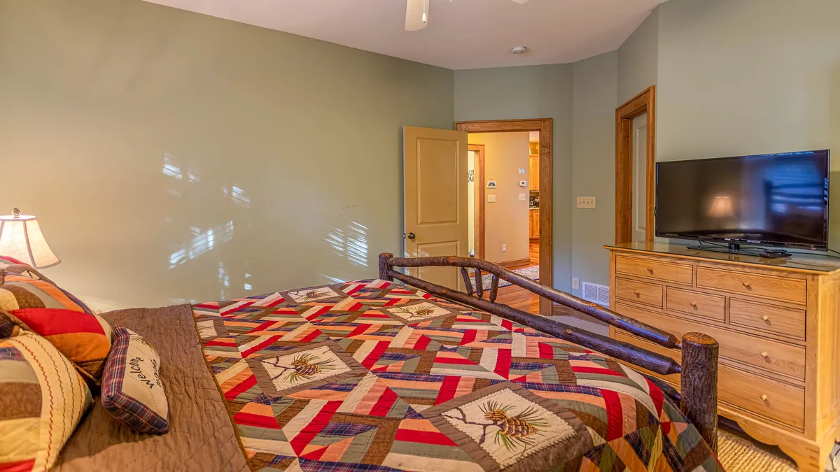 A comfortable bedroom with a bed draped in a colorful quilt, a wooden dresser, and a TV. The room feels inviting, with plenty of natural light and a doorway leading to the kitchen, suggesting easy access to amenities.