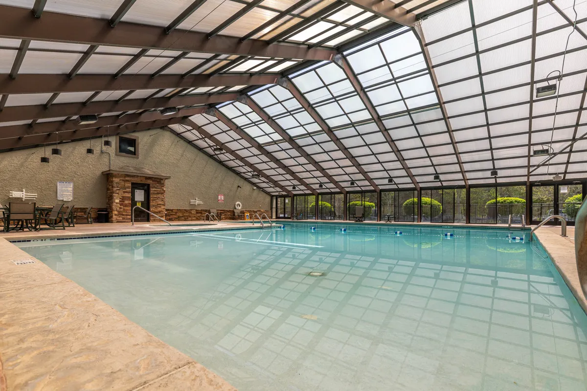 A spacious indoor pool room with a glass roof, plush lounge chairs, and a cozy stone fireplace. Soft daylight fills the space, enticing guests to enjoy swimming or relaxing poolside regardless of the weather.