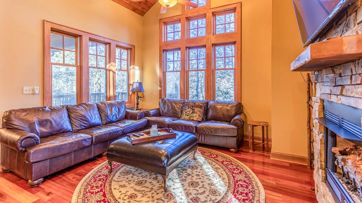 This image showcases a grand great room with a large leather sectional sofa set around a dramatic stacked-stone fireplace. Expansive windows line the wall, filling the space with natural light and offering stunning mountain views. An ornate area rug and rustic accents complete the inviting ambiance, while the open concept allows for easy flow between seating areas.