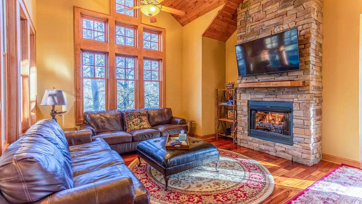 A spacious living area featuring large windows that flood the room with natural light, a stone fireplace as a central gathering point, plush leather sofas, a decorative rug, and a wall-mounted TV. The inviting setup is ideal for family gatherings, watching movies, or relaxing by the fire.
