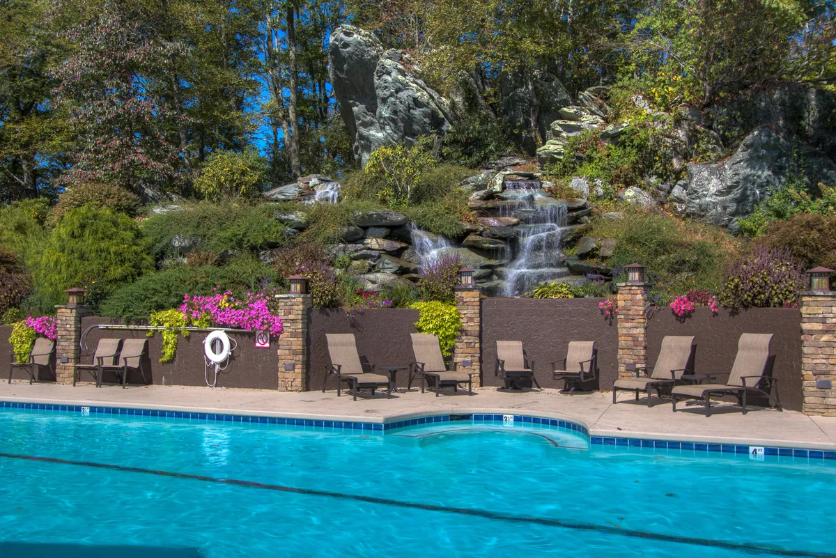 An exterior view of the resort's swimming pool, bordered by a landscaped garden and shaded by surrounding greenery. Deck chairs line the pool, creating a sense of leisure and community. The area is bright and peaceful, evoking the feeling of a sophisticated mountain escape.